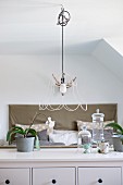 Pendant lamp with wire frame lampshade above orchids and glass ornaments on sideboard; double bed in background