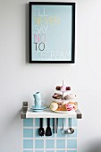 Pastries on cake stand and espresso cups on bracket shelf with kitchen utensils hung below under framed poster with motto