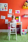 Ausschnitt eines farbigen Kinderzimmers, Kind auf hohem, weißem Stuhl vor Schreibtisch, an orangeroter Wand Kinderzeichnungen