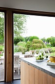 Glass bowls of apples on stone worksurface of island counter; glass wall with view of terrace and large garden