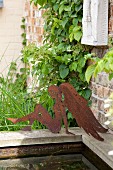 Rusty, metal angel silhouette on edge of partially visible pool in garden