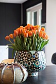 Üppiger, orangefarbener Tulpenstrauss in Retro Vase mit geometrischem Muster auf Tisch