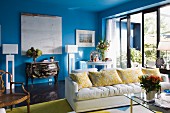 weiße Polstercouch neben Glas-Terrassentür, an blauer Wand Rokoko Kommode im Wohnzimmer