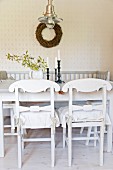 White-painted, wooden kitchen chairs with curved backrests at dining table in front of wicker wreath on wall