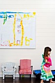Children's chairs below child's drawing on white wooden wall; little girl to one side