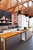 Narrow wooden counter with base units in open-plan, white designer kitchen with pendant lamps suspended from exposed wooden roof structure in modern country house