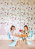 Little girls sitting on pale blue chairs at round table in front of fitted cupboards covered in butterfly-patterned wallpaper