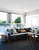 Grey corner sofa in front of glass wall with half-height folding windows and view of pool; Scandinavian stools used as side tables