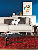 Schlichtes, beiges Sofa und Glastisch auf rot gemustertem Orientteppich; dekoratives Farbkreisbild vor blau getönter Wand