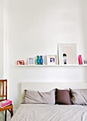 Row of pillows and cushions on bed below pictures and figurines on white floating shelf in bedroom