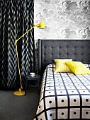 Double bed with grey upholstered headboard next to yellow, retro standard lamp against wall with cloud-patterned wallpaper