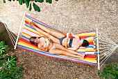 Two girls wearing bikinis relaxing in hammock whilst reading books