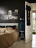 Bed with upholstered headboard below photos tacked on black-painted wall next to open interior door