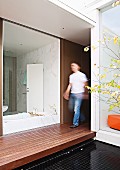 View from courtyard with wooden platform through frameless glass wall into white bathroom opposite home office; blurred man walking between the rooms