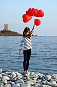 Little girl holding red, heart-shaped balloons on pebbly beach