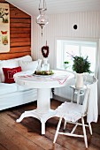 White, round dining table and chair, fir branches in jug on side table in front of window in wood-clad attic room