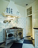 Vintage, stainless steel cooker under kitchen utensils suspended from extractor hood on tiled wall and diagonal chequered floor