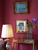 Vase of flowers next to candlesticks on vintage console table below gilt-framed pictures on claret wall; floor lamp with pale fabric lampshade