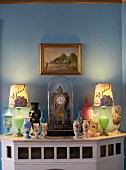 Various painted vases and table clock under glass cover on wood-clad fireplace below gilt-framed landscape on wall painted pale blue