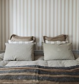 Stacked pillows in coordinated colours on French bed against wall with striped wallpaper in traditional bedroom with subdued colour scheme