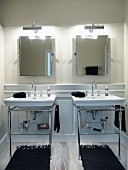 Twin vintage washstands on metal frames below two illuminated mirrors on wall with black rugs on white wooden floor