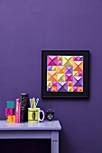 Hand-crafted, 3D-effect artwork made from blue, green and purple origami squares of different sizes on violet wall; matching ornaments on desk