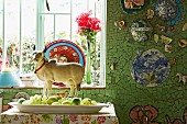 Animal figurine behind dish of various green fruits below window in wall tiled in mosaic of green tile fragments