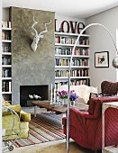 Red armchair below retro arc lamp and rustic coffee table in lounge area opposite open fireplace below stylised hunting trophy on grey rendered wall