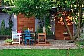Verschiedenfarbige Stühle und Sitzbank auf gekiestem Terrassenplatz mit begrüntem Sichtschutz an sommerlichem Tag
