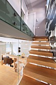Staircase with pale, solid wood treads, glass partition and view into interior with open-plan kitchen area