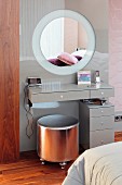 Minimalist dressing table and silver-coloured stool in front of pale grey glass panel with integrated round mirror