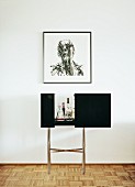 Minibar with black sliding doors on chrome frame below contemporary drawing on wall