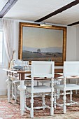 White-framed dining chairs with turned legs around dining table below gilt-framed landscape painting