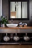 Modern washstand with white basin, three metal vases on shelf below and framed mirror on grey, marbled wall