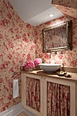 Illuminated washbasin on base unit with red and white patterned fabric panels in doors and matching wallpaper in attic room