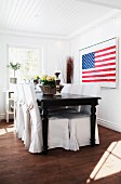 Stars and Stripes artwork in dining area with black, antique wooden table and chairs with loose covers in white, country-house interior