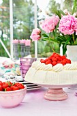 Strawberry cream cake on cake stand, bowl of strawberries and peonies on table in conservatory