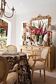 Rococo-style chairs in elegant dining area with vase of gladioli and table lamps on pale wooden cabinet