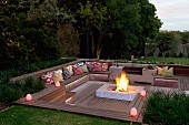 Abendstimmung über elegantem Sitzplatz im Garten, tiefer gelegte Holzterrasse mit integrierten und Sitzbänken, in der Mitte grosse Feuerstelle mit Feuer