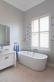 Free-standing bathtub below window with closed interior shutters next to white, rustic washstand
