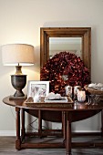Wooden console table decorated with festive wreath of red leaves, tealight holders and table lamp with pale fabric lampshade