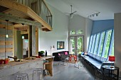 Offenes Wohnzimmer mit rustikalem Kücheneinbau unter Galerie auf Holzkonstruktion, gegenüber gekippte Fensterfront in Hellblau