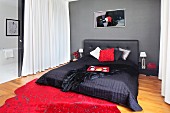 Black double bed with upholstered headboard and blanket against grey wall; red, speckled rug on floor