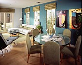 Graue, gepolsterte Stühle an rundem Glastisch, dahinter gemütliche Sofaecke in offenem Wohnraum mit modernen Bildern an blauer Wand
