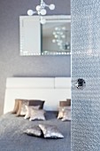 A glass door with a knob in the foreground and an unfocussed view into a bedroom with a double bed, silver decorative cushions and a grey quilt