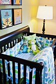 Schwarzes Babybett mit blau-grüner Bettwäsche und Stofftieren in Zimmerecke mit Stehlampe & lustigen Wandbildern