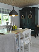 Simple dining table and white kitchen chairs below vintage pendant lamp; farmhouse cupboard painted black with wreaths on doors