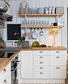 White, country-house-style kitchen; table lamp on kitchen counter below shelves of glasses and mugs on wooden wall