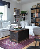 Lit candles in white candlesticks on rustic wooden trunk in front of white sofa and easy chair in corner of living room