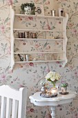 Vase of flowers on side table below shelving on wall with floral wallpaper